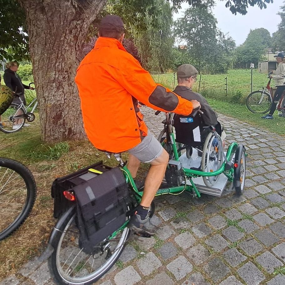 Rollstuhlfahrrad über Aktion Mensch gefördert worden Ein Radfahrer mit orangefarbener Jacke schiebt einen Passagier auf einem speziellen Fahrrad.